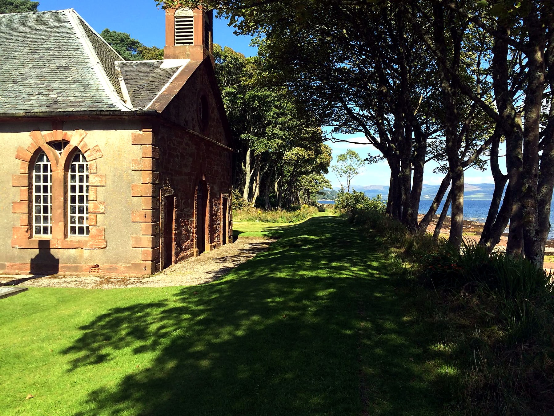 A small harled chapel with red sandstone windows and a Celtic cross beside the shore path on the Mount Stuart estate, Isle of Bute